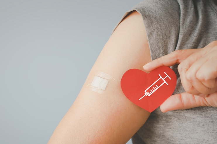 Senior woman holding red heart shape with syringe and showing her arm with bandage